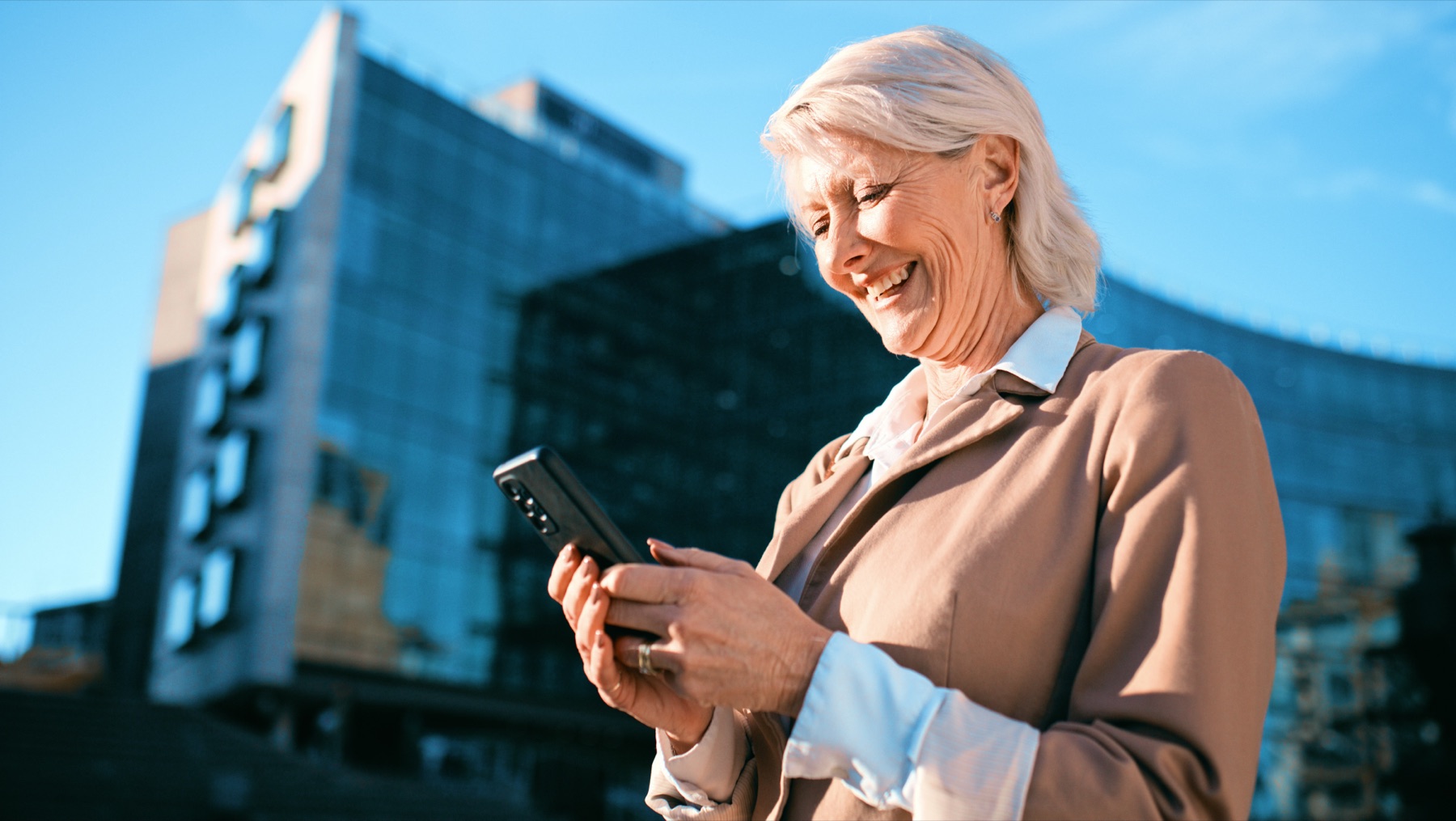 A woman smiling while looking at her phone, city building and blue sky behind her